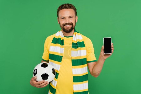 cheerful man in striped scarf holding football and smartphone with blank screen isolated on green