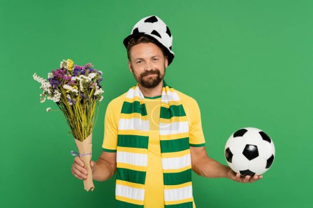 Téléchargez les photos : Homme barbu en chapeau de ventilateur de sport tenant le football et les fleurs sauvages isolés sur vert - en image libre de droit