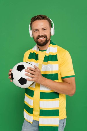 fanático de los deportes barbudos felices en bufanda y auriculares que sostienen el fútbol aislado en verde