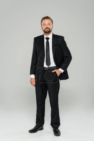 Foto de Longitud completa del hombre barbudo en ropa formal de pie con la mano en el bolsillo en gris - Imagen libre de derechos