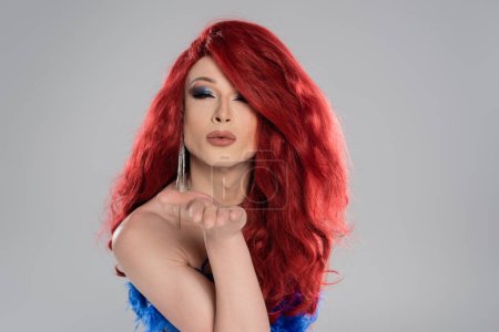 Retrato de elegante drag queen en peluca roja soplando aire beso aislado en gris  