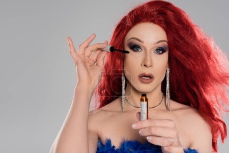 Retrato de elegante drag queen en peluca roja sosteniendo rímel y mirando a la cámara aislada en gris