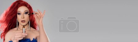 Téléchargez les photos : Drag queen en perruque rouge tenant le mascara et regardant la caméra isolée sur gris, bannière - en image libre de droit