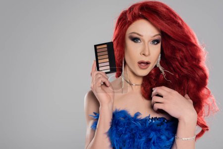 Transgender person in red wig holding eye shadows isolated on grey  