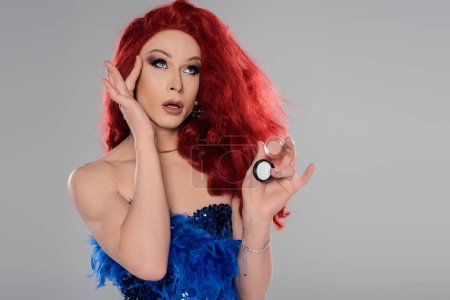 Stylish drag queen holding concealer and touching face isolated on grey  