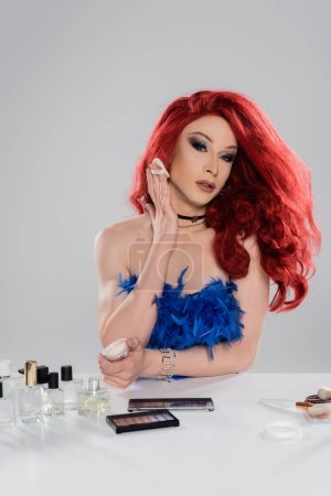 Portrait of drag queen holding face powder near perfumes and decorative cosmetics isolated on grey  