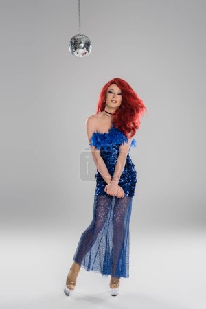 Smiling drag queen in dress and heels looking at camera near disco ball on grey background 