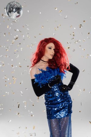 Glamorous drag queen in dress and gloves standing under confetti and disco ball on grey background 