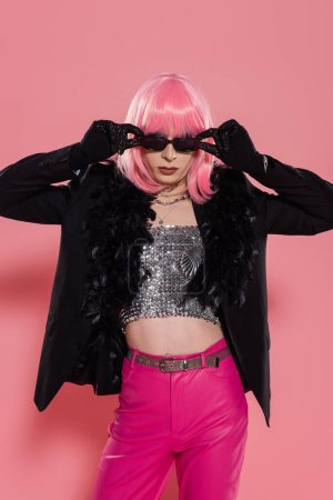 Stylish drag queen in jacket with feathers holding sunglasses on pink background 