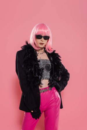 Foto de Trendy drag queen en gafas de sol y chaqueta con plumas de pie sobre fondo rosa - Imagen libre de derechos