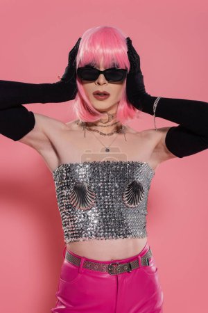 Trendy drag queen in sunglasses and gloves touching wig on pink background 