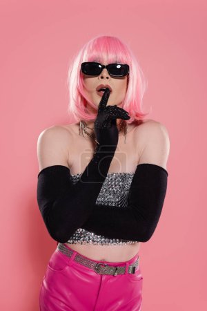 Stylish drag queen in sunglasses and gloves showing hush gesture on pink background 