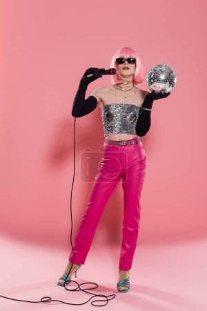 Full length of trendy drag queen in sunglasses and gloves holding microphone and disco ball on pink background 