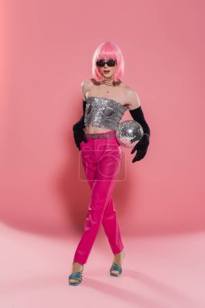 Trendy drag queen in sunglasses and shiny top posing with disco ball on pink background 