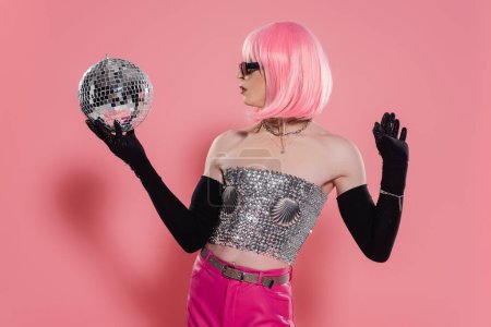 Foto de Vista lateral de la drag queen de moda en la parte superior brillante y guantes que sostienen la bola disco sobre fondo rosa - Imagen libre de derechos