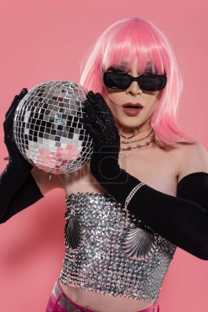 Portrait of stylish drag queen in sunglasses holding disco ball on pink background 