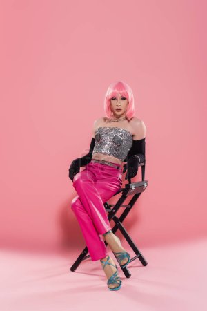 Fashionable drag queen in top and gloves posing while sitting on chair on pink background 