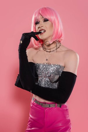 Portrait of stylish drag queen in gloves biting finger on pink background 