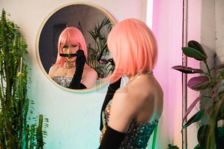 Blurred drag queen in wig wearing sunglasses near mirror at home 