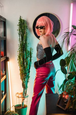Fashionable drag queen in sunglasses and gloves looking at camera near plants at home 