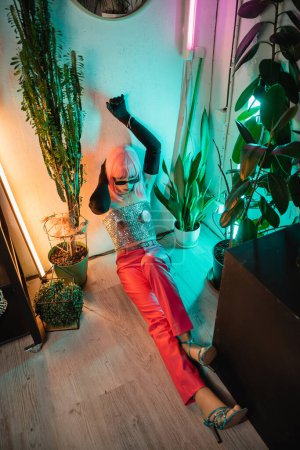 Hochwinkel-Ansicht des modischen Mannes mit Handschuhen und glänzendem Top, der auf dem Boden in der Nähe von Pflanzen zu Hause sitzt 