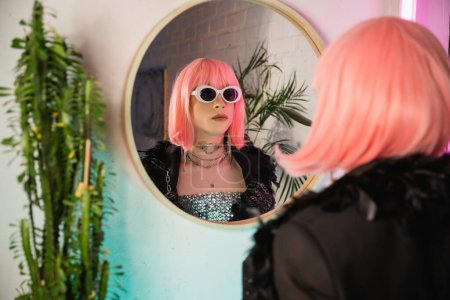 Fashionable drag queen in wig and sunglasses standing near mirror at home 