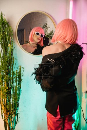 Foto de Persona transexual de moda en chaqueta y peluca con gafas de sol cerca del espejo en casa - Imagen libre de derechos