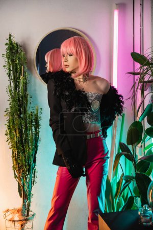 Trendy drag queen en peluca rosa y chaqueta de pie cerca de las plantas en casa 