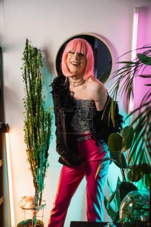 Carefree drag queen in pink wig looking at camera near plants at home 