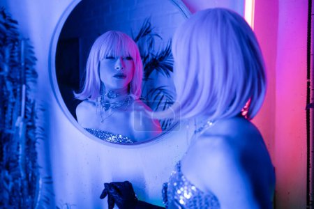 Blurred drag queen in wig and top looking at mirror in neon light at home 