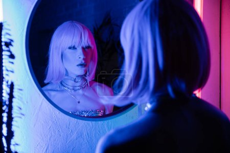 Elegante drag queen en peluca mirando el espejo cerca de la luz de neón en casa 