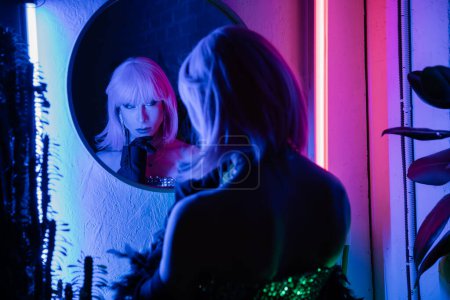 Modische Drag Queen schaut in den Spiegel, während sie zu Hause neben Pflanzen und Neonlicht steht  