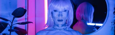 Portrait of drag queen in wig looking at camera in neon lighting at home, banner 