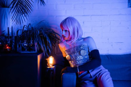 Drag queen in wig and top looking at lamp near plants at home 