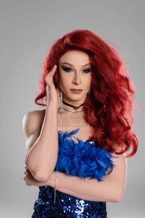 Drag queen in red wig and dress posing and looking at camera isolated on grey 