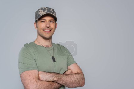 soldado estadounidense feliz en camiseta y gorra militar de pie con los brazos cruzados durante el día conmemorativo aislado en gris