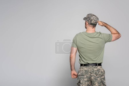 vista trasera del militar en uniforme militar y gorra saludando sobre fondo gris 