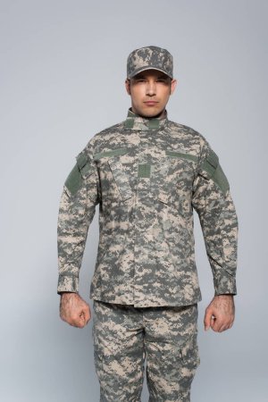 Foto de Soldado patriótico en uniforme militar y gorra de pie con los puños cerrados aislados en gris - Imagen libre de derechos