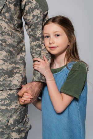 linda niña preadolescente abrazando la mano del padre en uniforme militar aislado en gris 