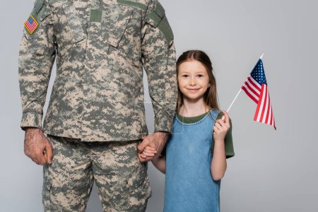 Foto de Alegre niña preadolescente sosteniendo la bandera americana y la mano del padre en uniforme militar durante el día conmemorativo aislado en gris - Imagen libre de derechos