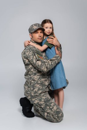 preteen girl hugging father in military uniform and cap during memorial day on grey 