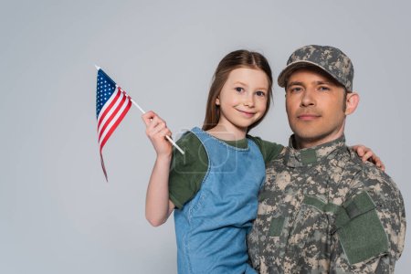serviceman in military uniform holding in arms happy daughter with American flag isolated on grey 