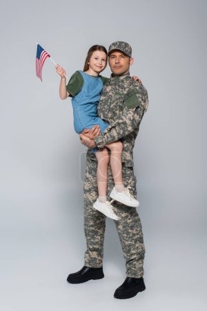 Téléchargez les photos : Militaire en uniforme militaire tenant dans les bras fille joyeuse avec drapeau américain sur gris - en image libre de droit