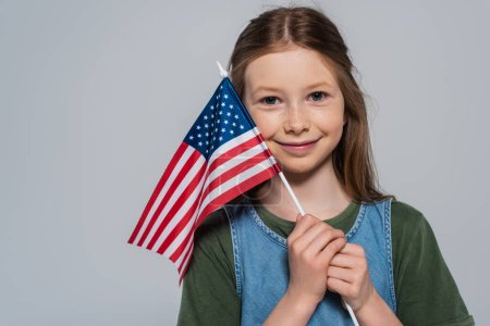 niña alegre y patriótica sosteniendo la bandera de Estados Unidos de América durante el día conmemorativo aislado en gris 