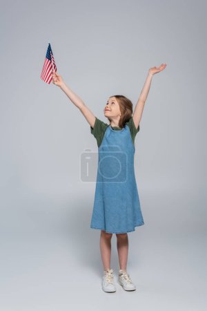 Foto de Longitud completa de niña preadolescente patriótica sonriendo mientras sostiene la bandera de Estados Unidos de América durante el día memorial en gris - Imagen libre de derechos