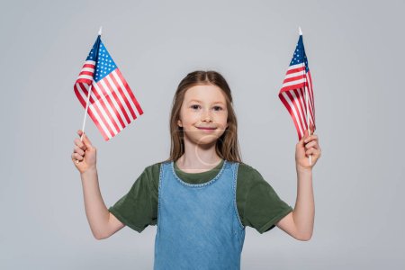 Patriotische preteen girl lächelnd, während sie Flaggen von Amerika während des Gedenktages isoliert auf grau 