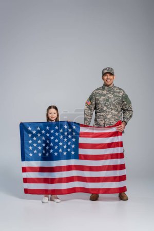 Foto de Militar con uniforme militar sosteniendo bandera de Estados Unidos con hija feliz en gris - Imagen libre de derechos