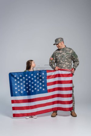 Foto de Alegre militar en uniforme militar sosteniendo bandera de Estados Unidos con hija feliz en gris - Imagen libre de derechos