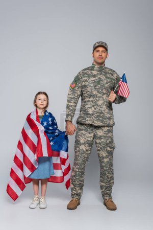 Foto de Militar con uniforme militar que sostiene la bandera de EE.UU. y la mano de hija feliz en gris - Imagen libre de derechos