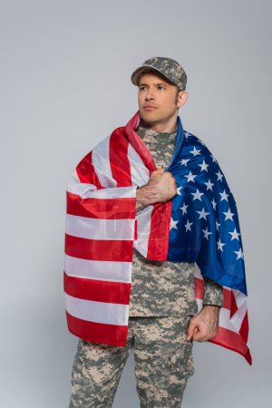 Patriotischer Armeesoldat in Tarnuniform, eingehüllt in die Flagge der Vereinigten Staaten von Amerika am Gedenktag isoliert auf grau 